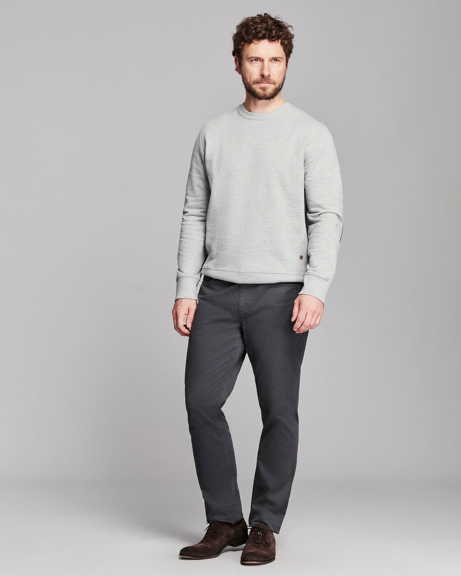 A man wearing a light gray long-sleeve crewneck sweater, dark gray trousers, and brown suede shoes, standing against a solid gray studio background.