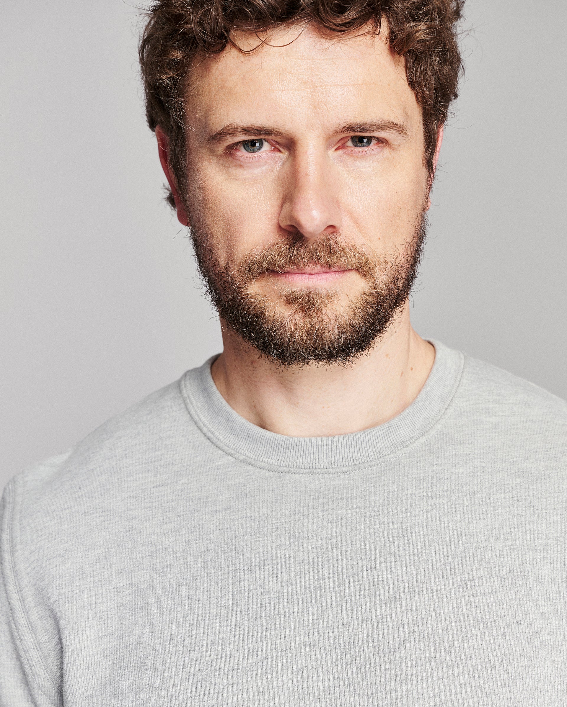 A close-up portrait of a man with curly brown hair and a beard, wearing a light gray crewneck sweatshirt against a solid gray background.
