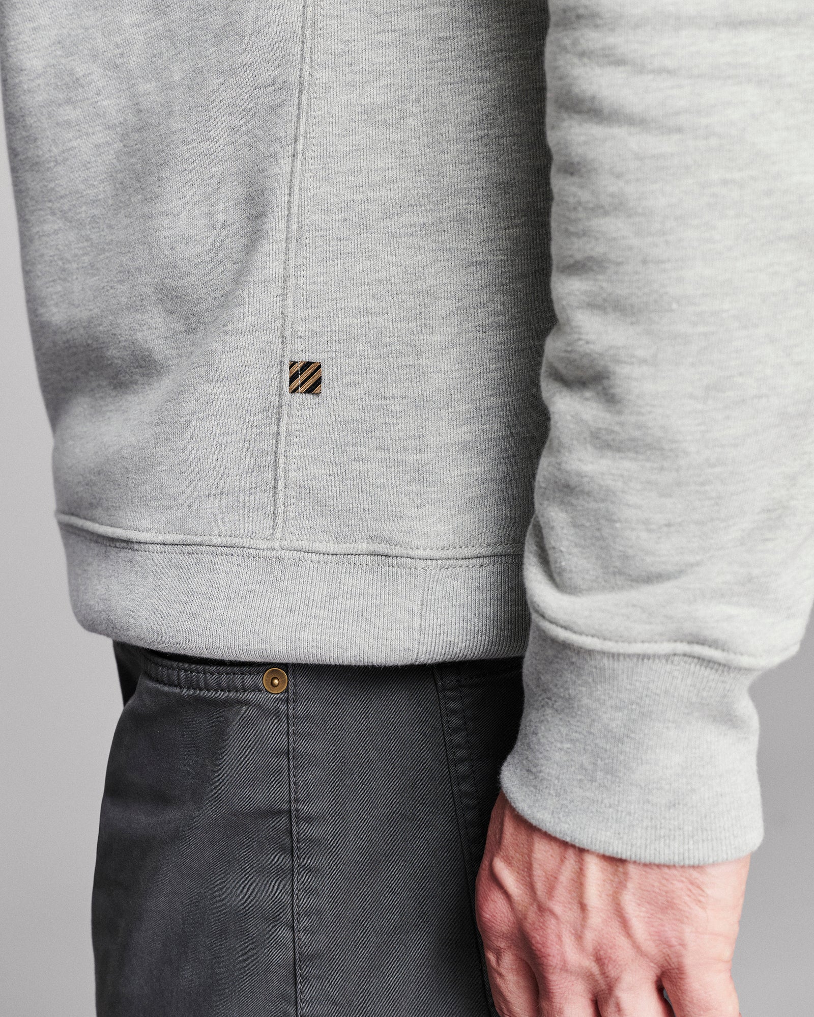 Close-up of a person wearing a light heather gray pullover sweatshirt with a small striped square logo patch on the chest, paired with dark gray pants against a neutral gray background.