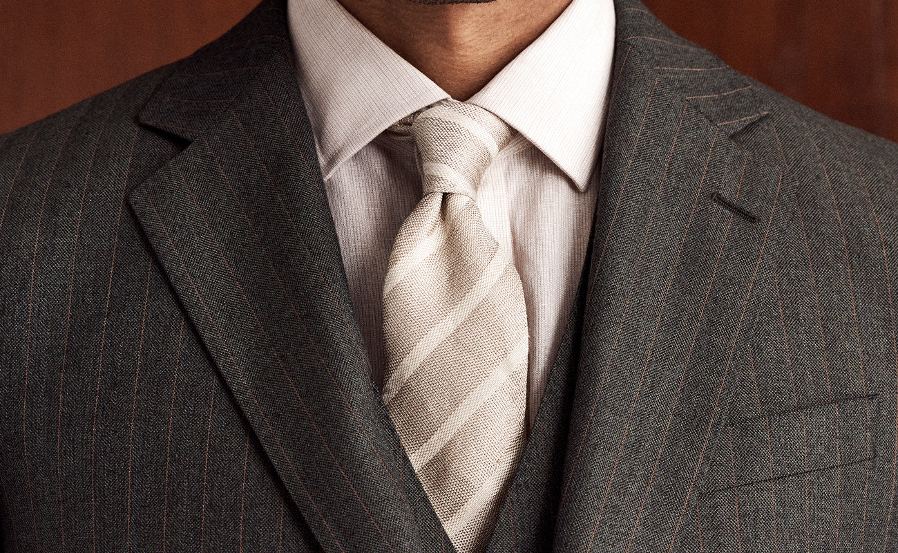 A close-up of a man wearing a dark gray pinstripe suit jacket, a light beige dress shirt, and a matching beige and white striped tie.