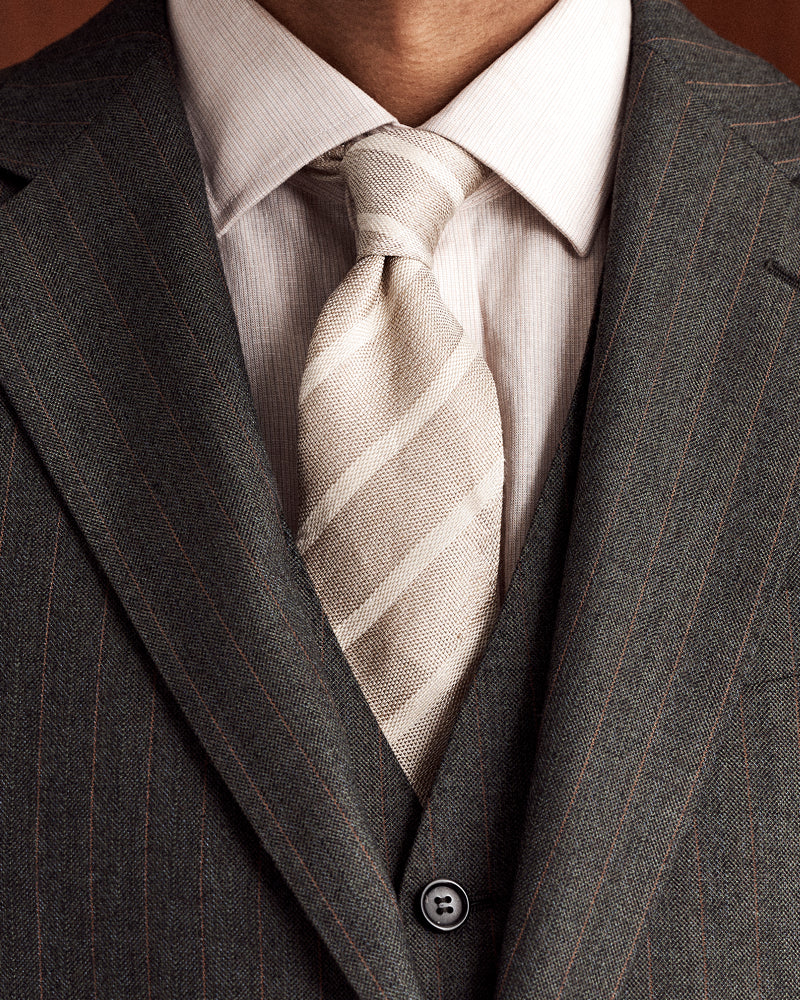 A close-up of a man's neck and chest wearing a dark gray pinstripe suit jacket, a matching vest, a white dress shirt, and a light beige striped tie.