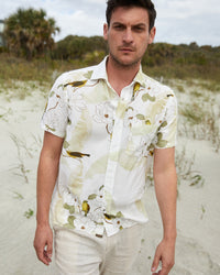 A man in a white floral short-sleeve shirt and light pants stands on a sandy beach with grass and trees in the background, looking directly at the camera.