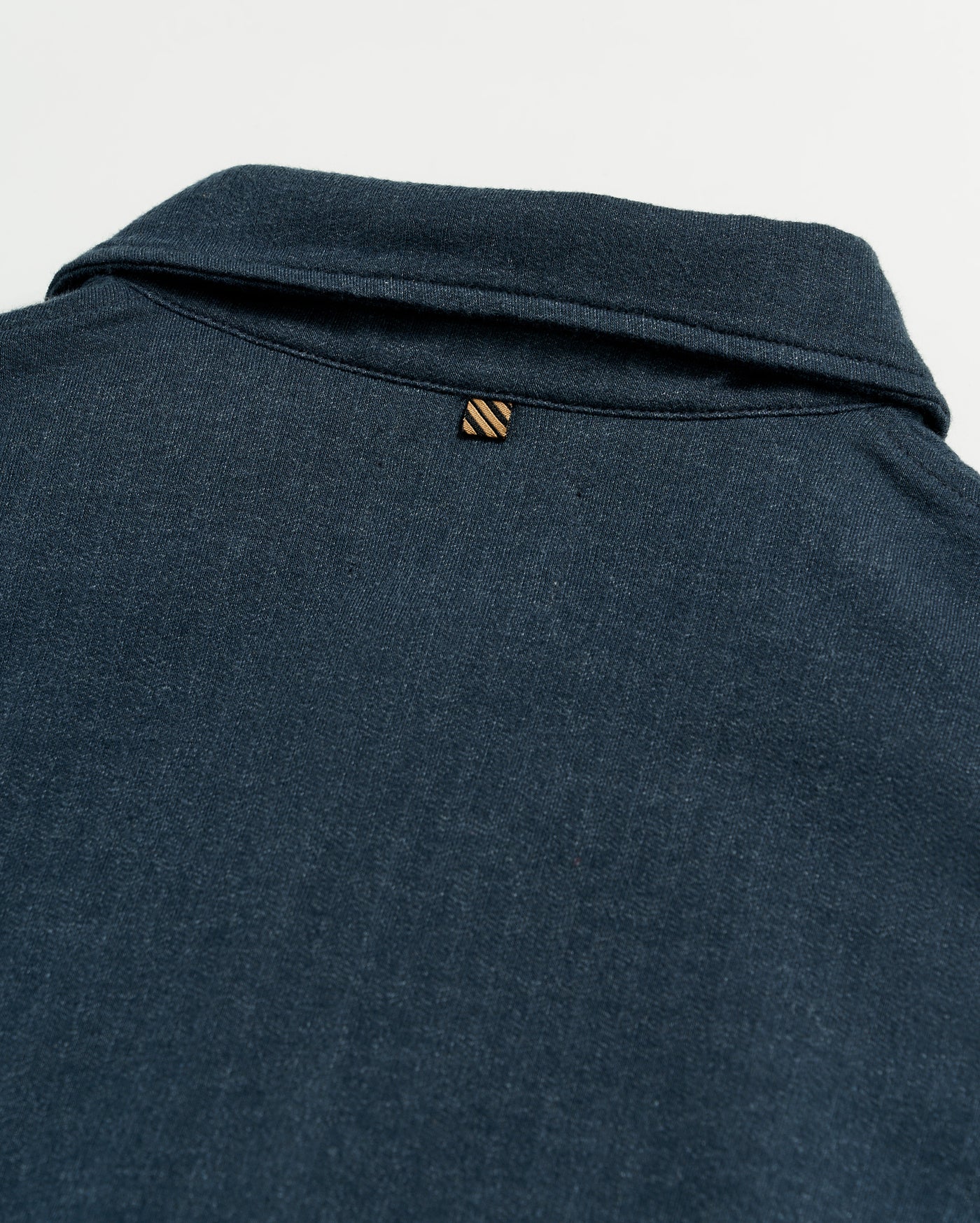 Close-up of the back of a dark blue collared shirt, showing a small, striped loop detail just below the collar on a plain white background.