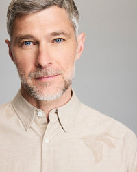 A middle-aged man with short gray hair and a trimmed beard wears a beige collared shirt with a subtle bird design. He is looking at the camera and smiling softly against a plain light background.