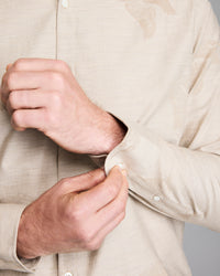 A person wearing a light beige, patterned shirt is buttoning the cuff of their sleeve with their right hand. The shirt has a subtle bird design. Only the hands and torso are visible.