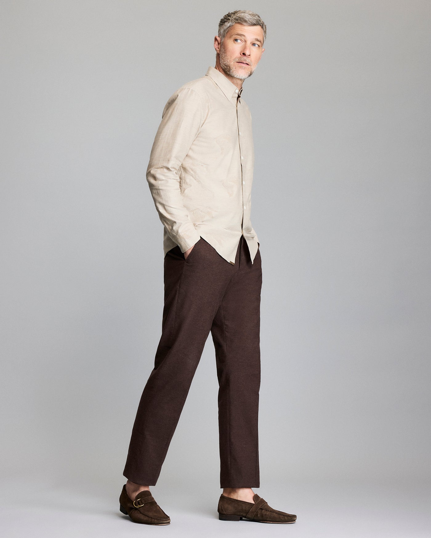 A man stands against a light gray background, wearing a beige button-up shirt, dark brown pants, and brown loafers. He has short, light hair and a beard, with hands in his pockets, gazing slightly to the side.