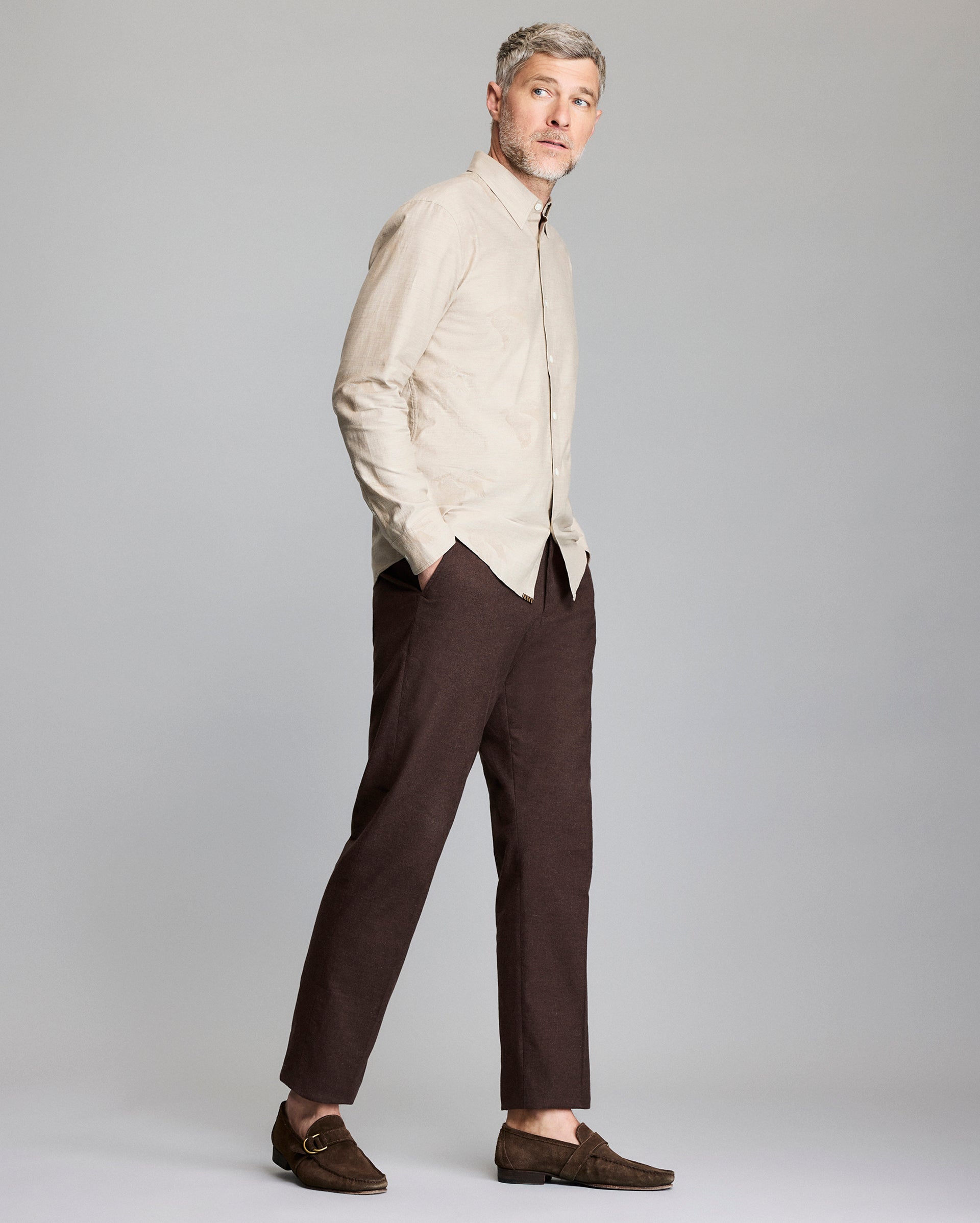 A man stands against a light gray background, wearing a beige button-up shirt, dark brown pants, and brown loafers. He has short, light hair and a beard, with hands in his pockets, gazing slightly to the side.