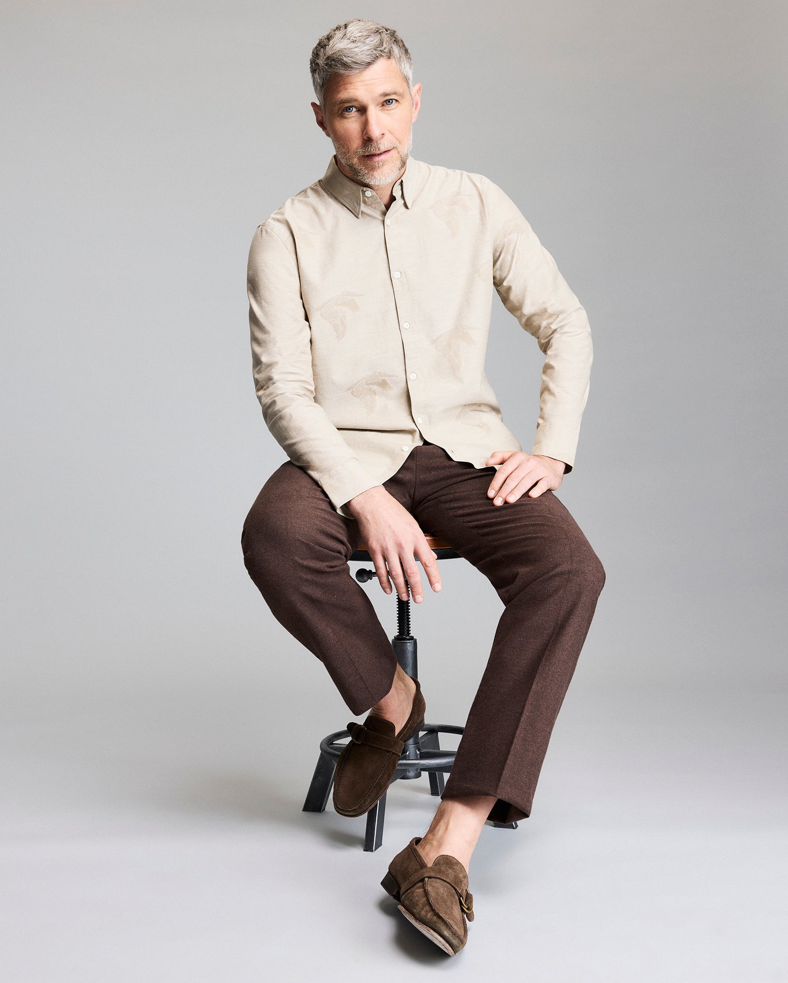 A man with short gray hair and a trimmed beard sits on a stool. He wears a beige button-up shirt, brown pants, and brown loafers. He looks at the camera against a plain light gray background.