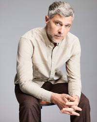 A middle-aged man with short gray hair and a beard sits on a stool, leaning forward with his hands clasped. He wears a beige button-up shirt and brown pants against a plain gray background.