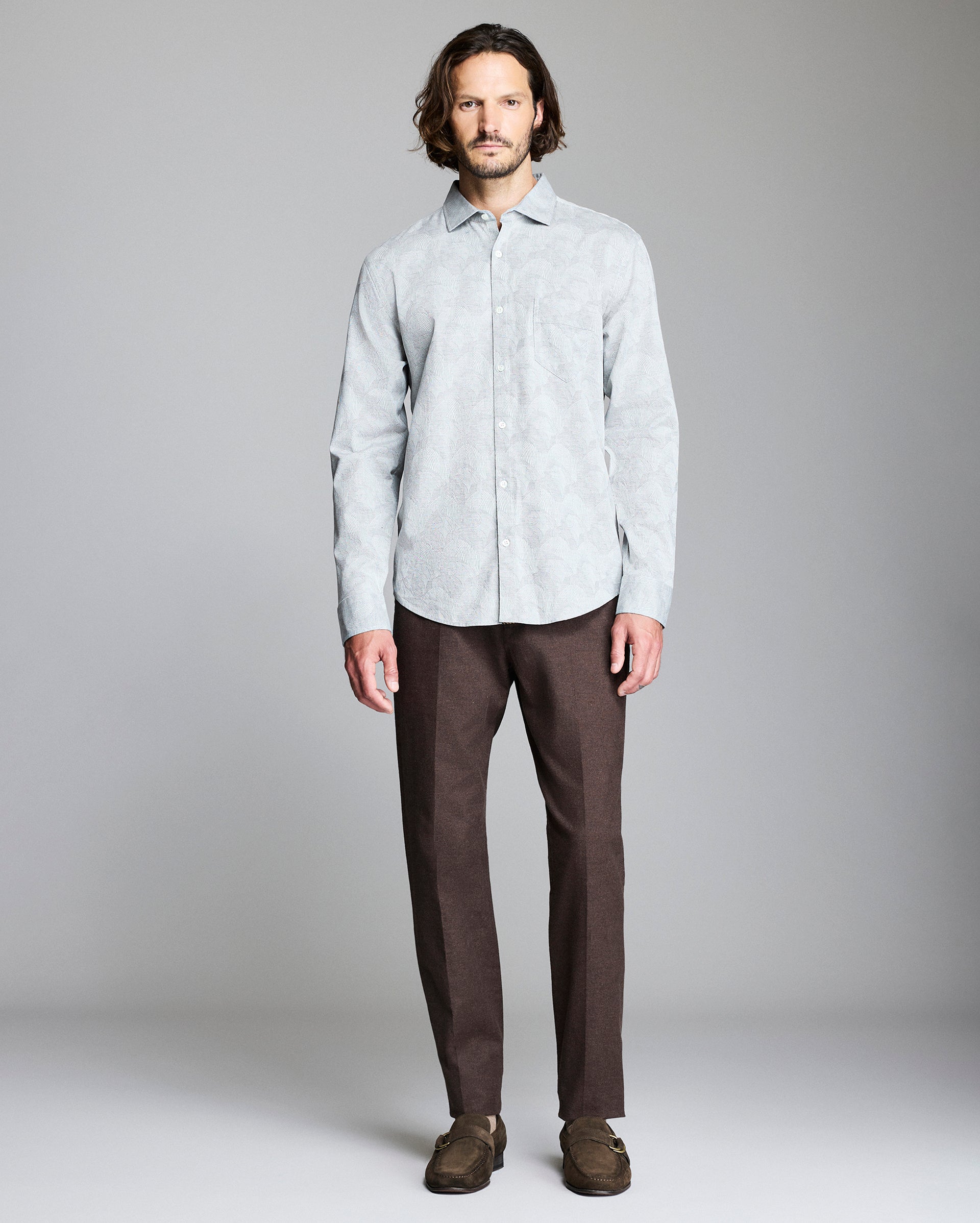 A man with wavy brown hair stands facing forward, wearing a light grey button-up shirt, brown pants, and brown loafers against a plain grey background.