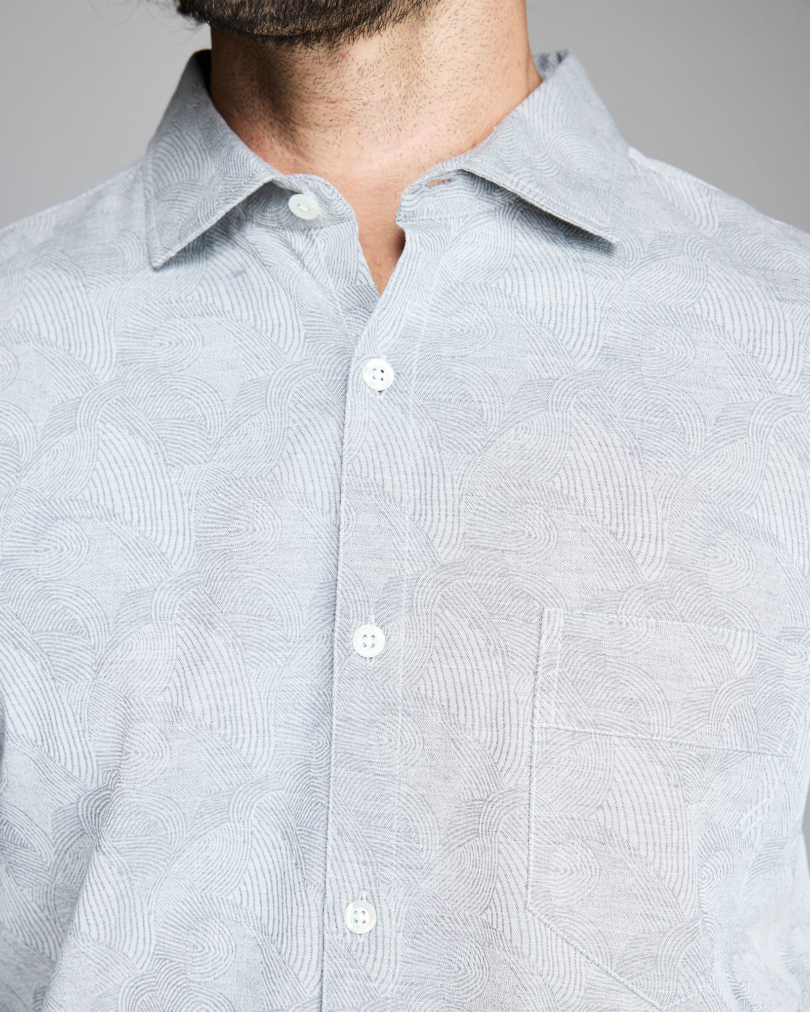 A close-up of a person wearing a light gray patterned button-up shirt with a collar and a chest pocket. The persons face is partially out of frame, showing only the lower part of their beard.