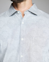 A close-up of a person wearing a light gray patterned button-up shirt with a collar and a chest pocket. The persons face is partially out of frame, showing only the lower part of their beard.