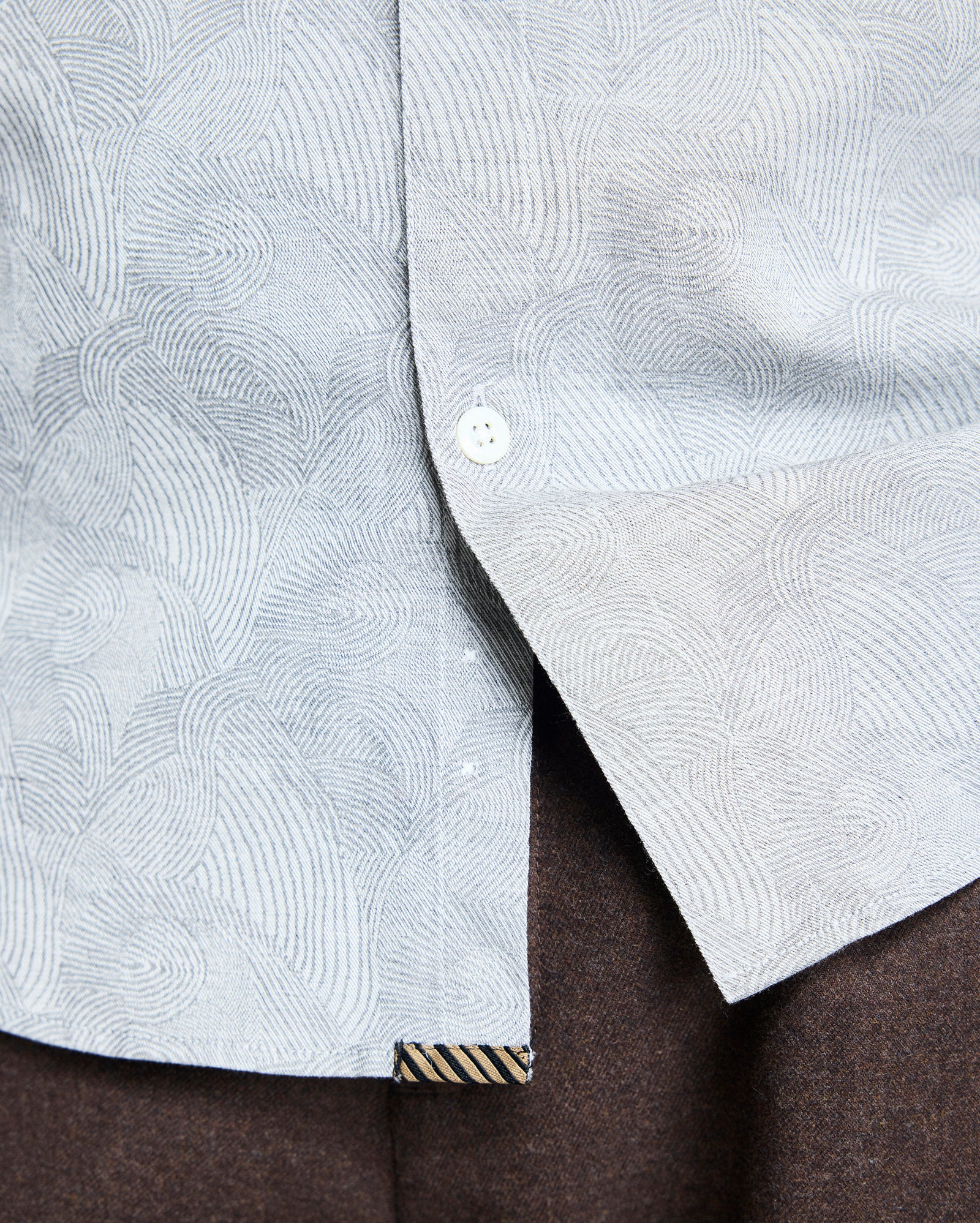 Close-up of a light gray patterned shirt with swirling lines, partially unbuttoned at the bottom, revealing dark brown pants underneath. A small striped fabric tag is visible near the shirt hem.