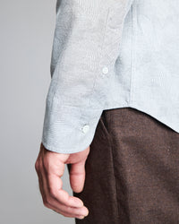 A close-up of a person’s arm and hand wearing a light blue patterned shirt with white buttons and brown pants, with the side seam and cuff of the shirt clearly visible.