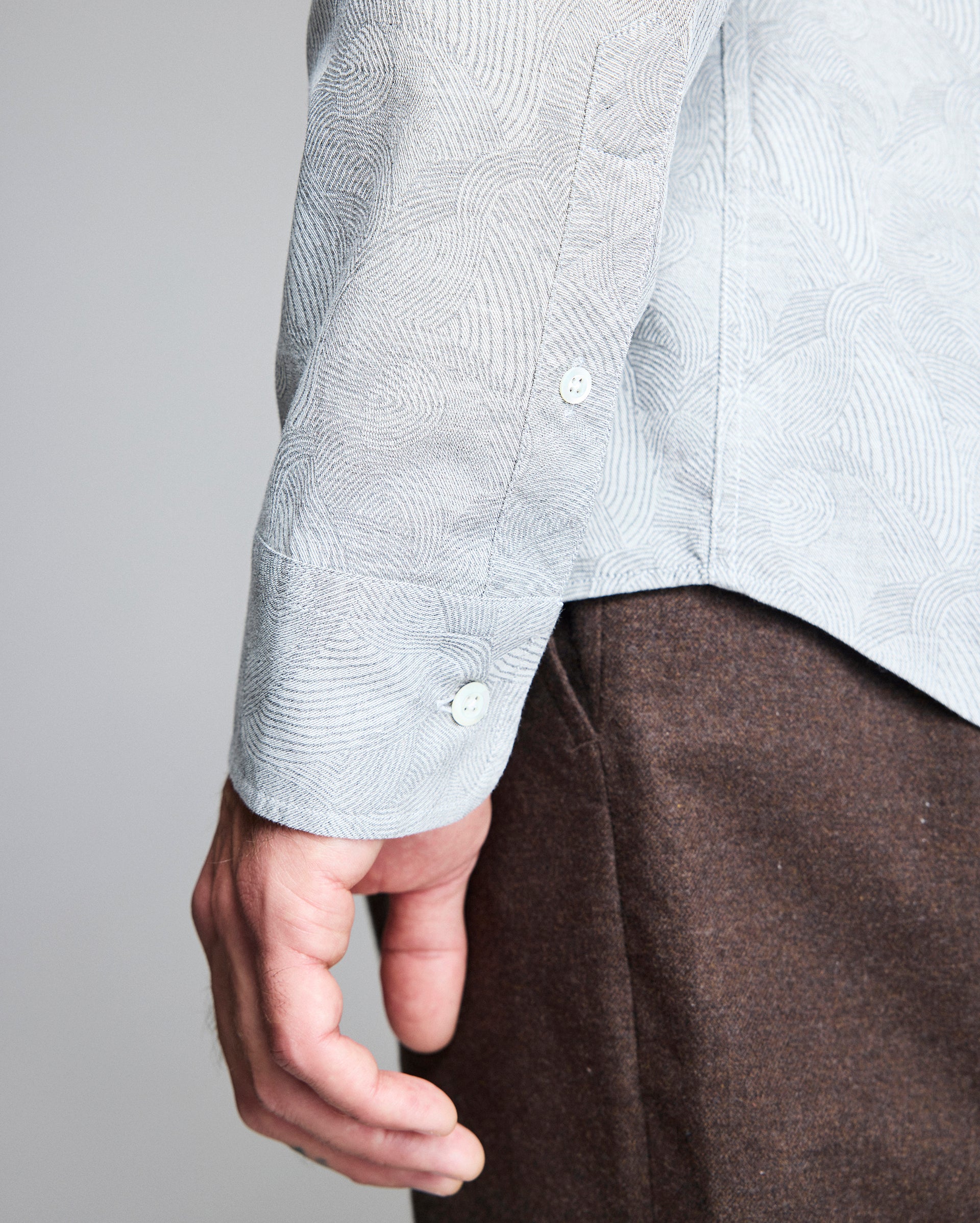 A close-up of a person’s arm and hand wearing a light blue patterned shirt with white buttons and brown pants, with the side seam and cuff of the shirt clearly visible.