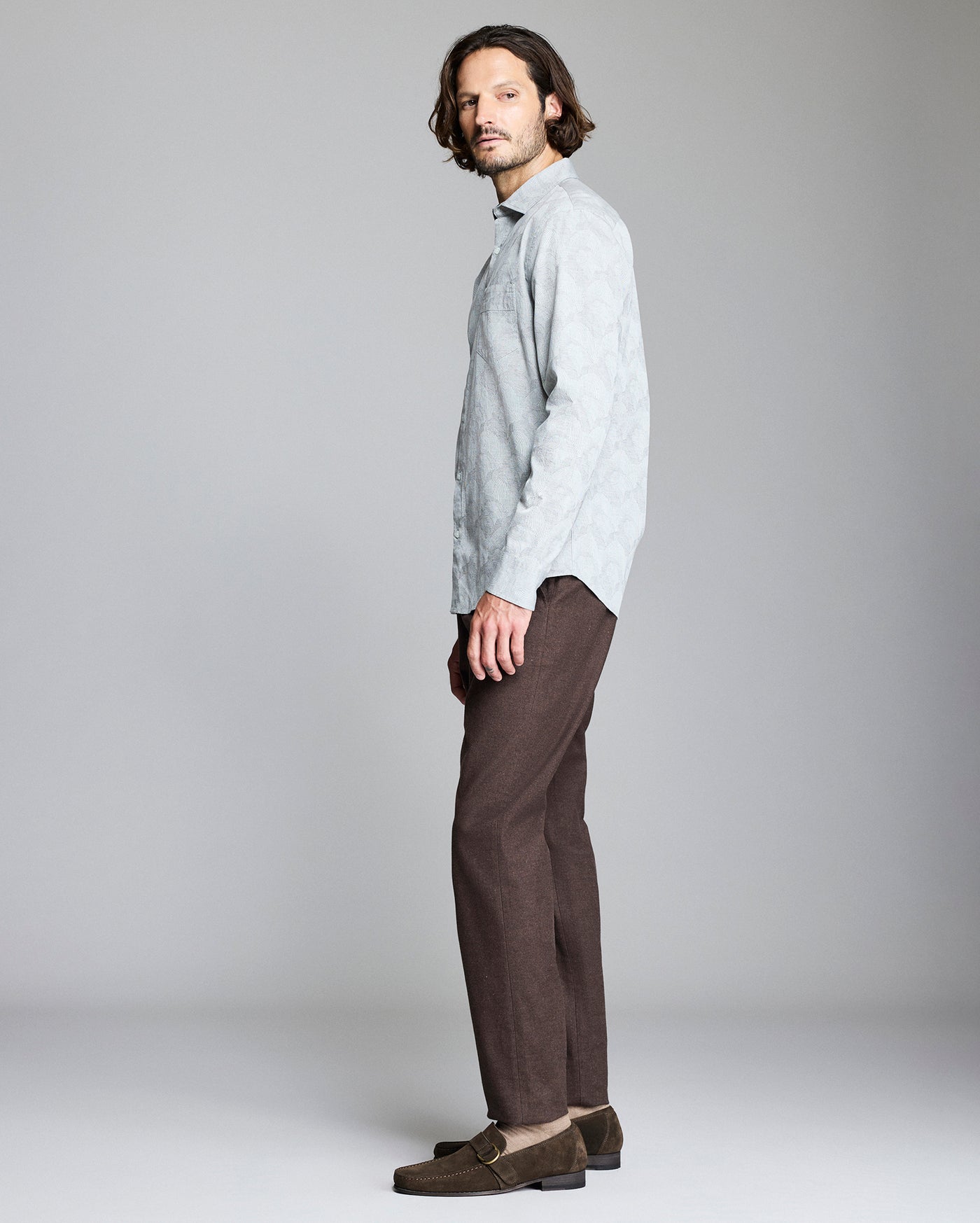 A man with medium-length brown hair, wearing a light gray long-sleeve shirt, brown pants, and brown loafers, stands sideways against a plain light gray background.