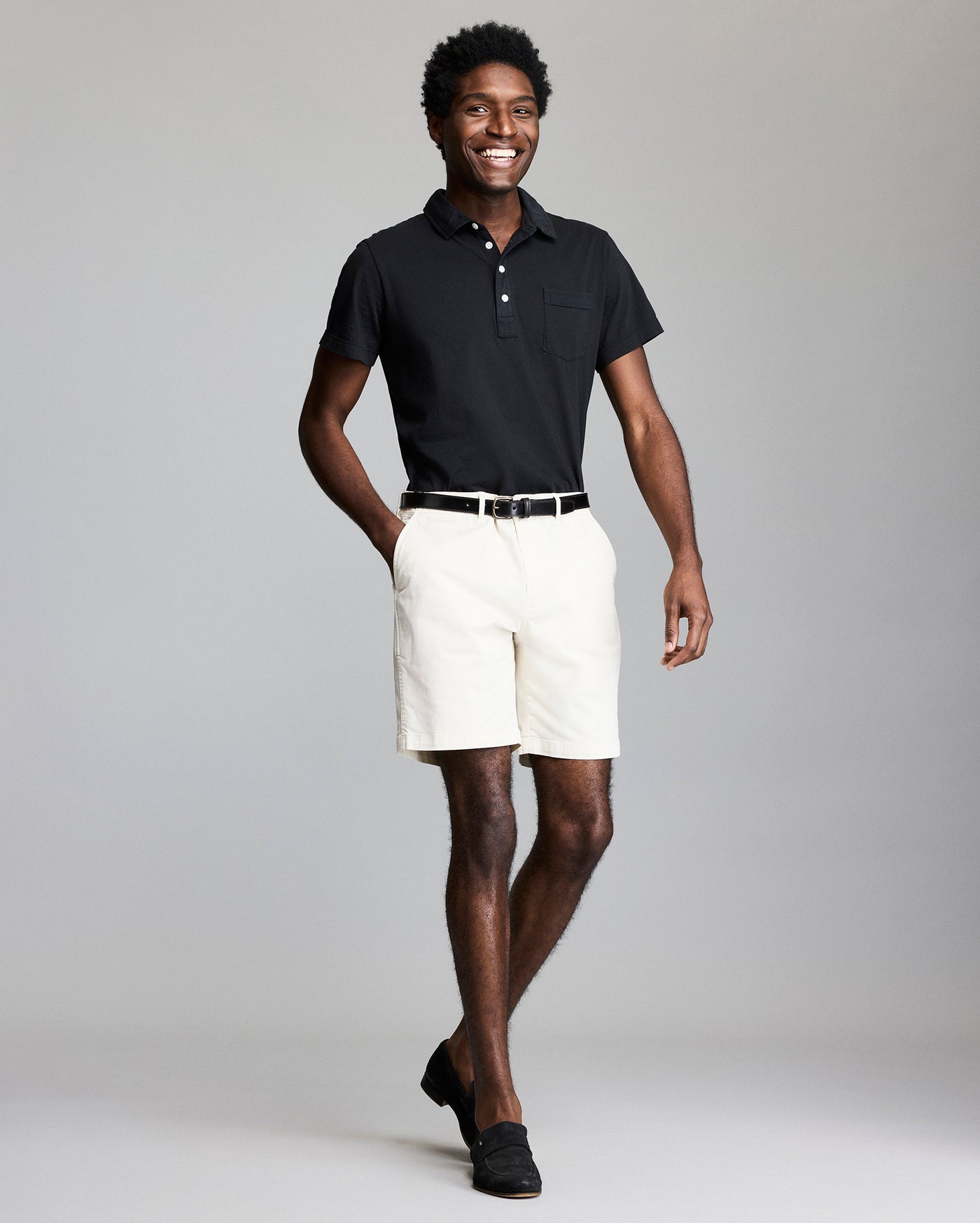 A smiling man stands against a plain gray background, wearing a black polo shirt, white shorts, a black belt, and black loafers. One hand is in his pocket and he appears relaxed and confident.