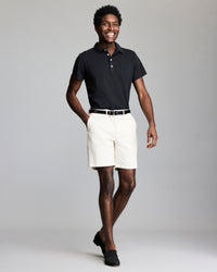 A smiling man stands against a plain gray background, wearing a black polo shirt, white shorts, a black belt, and black loafers. One hand is in his pocket and he appears relaxed and confident.