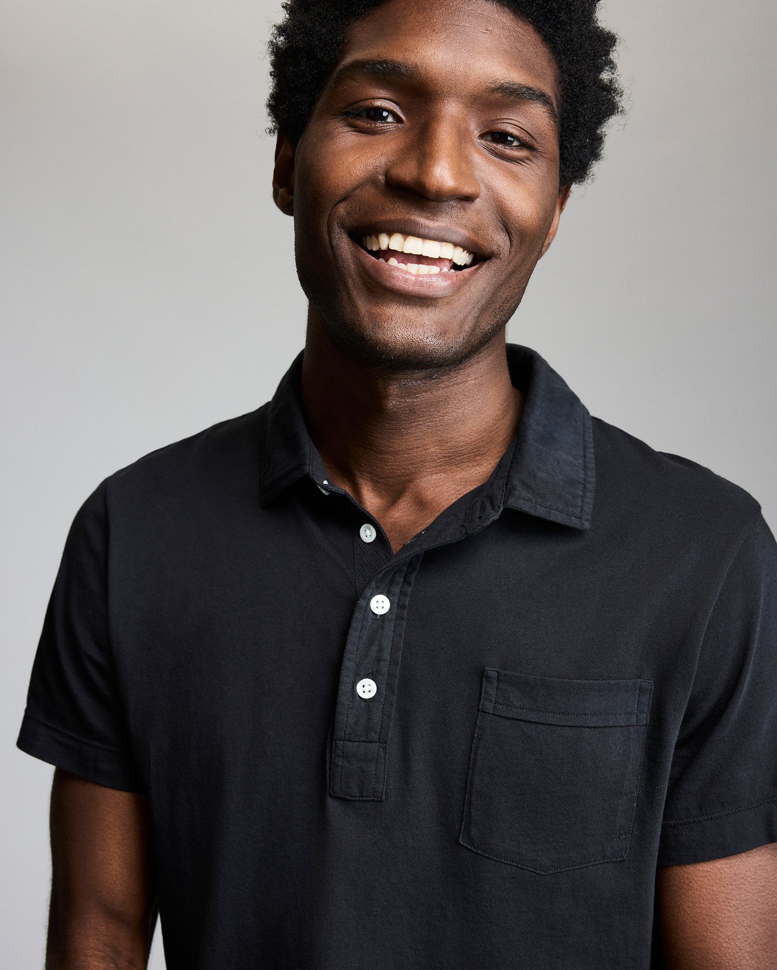 A man with short curly hair is smiling at the camera. He is wearing a black collared shirt with a front pocket against a plain, light background.