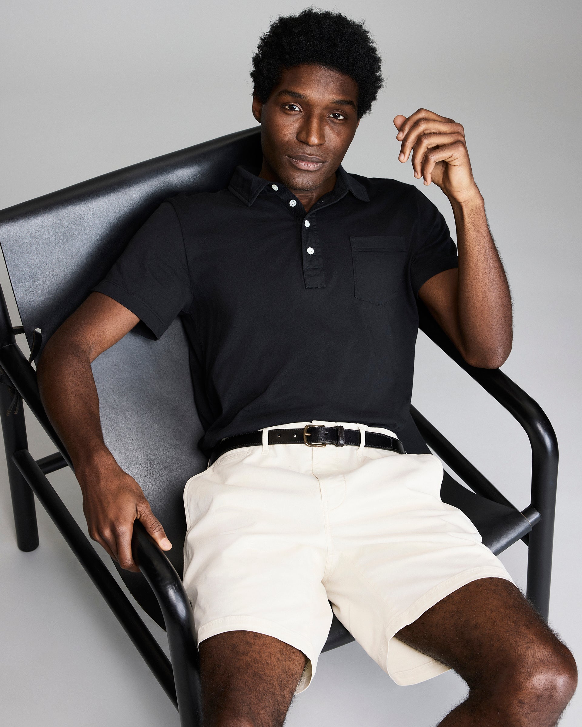 A man with short curly hair wears a black short-sleeve polo shirt, light-colored shorts, and a white belt, sitting relaxed in a modern black chair against a plain background.