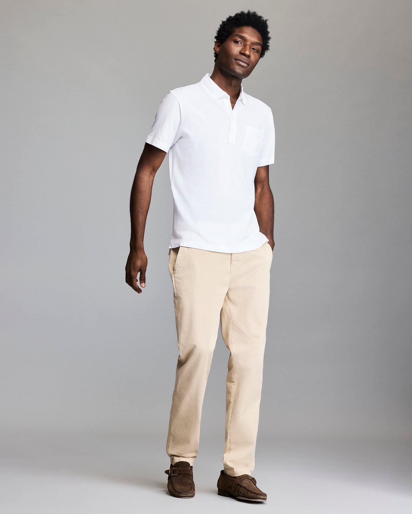A man stands against a plain gray background, wearing a white short-sleeve polo shirt, beige pants, and brown shoes. His left hand is in his pocket, and he is looking slightly to the side with a relaxed expression.