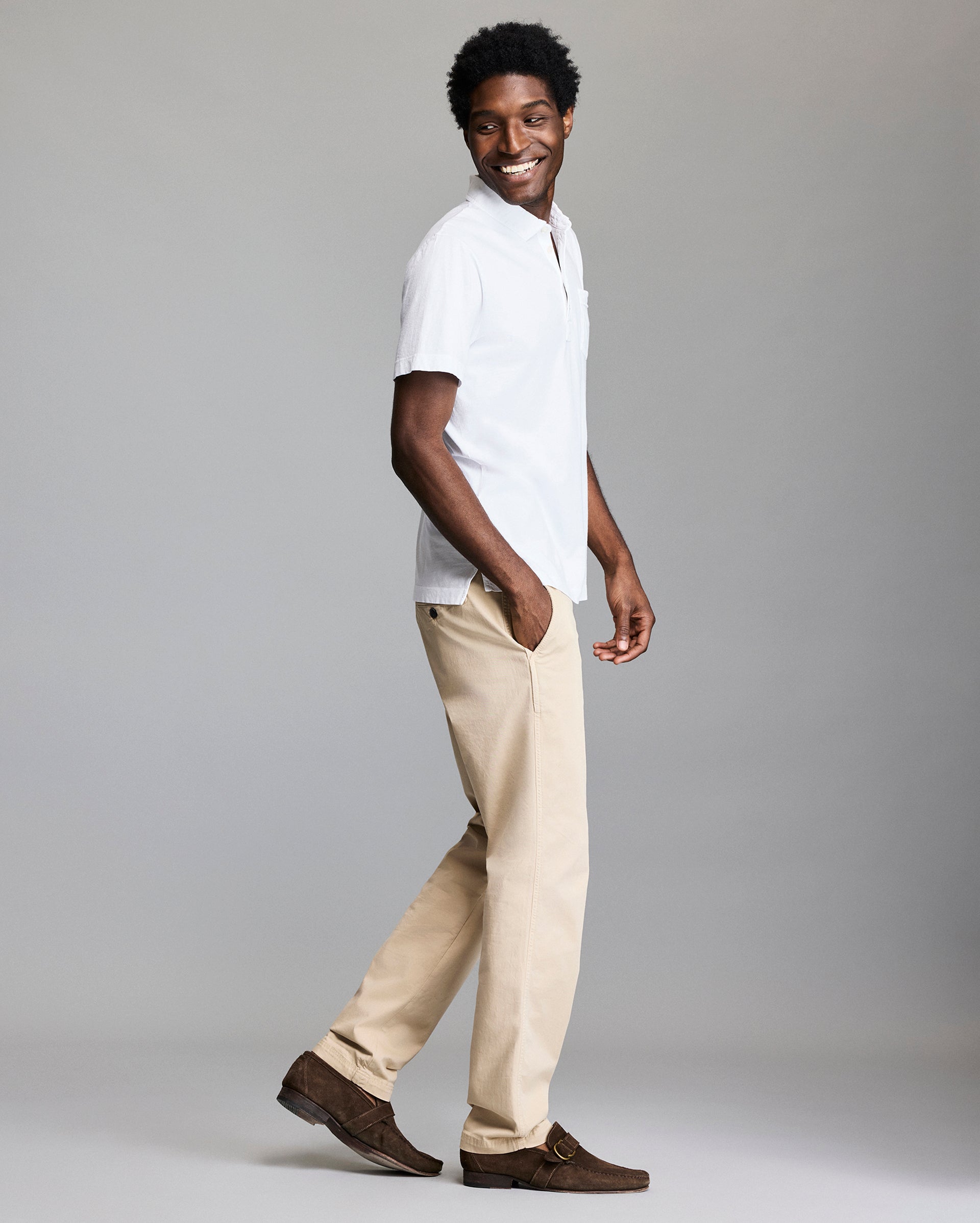A man stands sideways, smiling and looking over his shoulder. He is wearing a short-sleeved white shirt, beige pants, and brown shoes against a plain, light gray background.