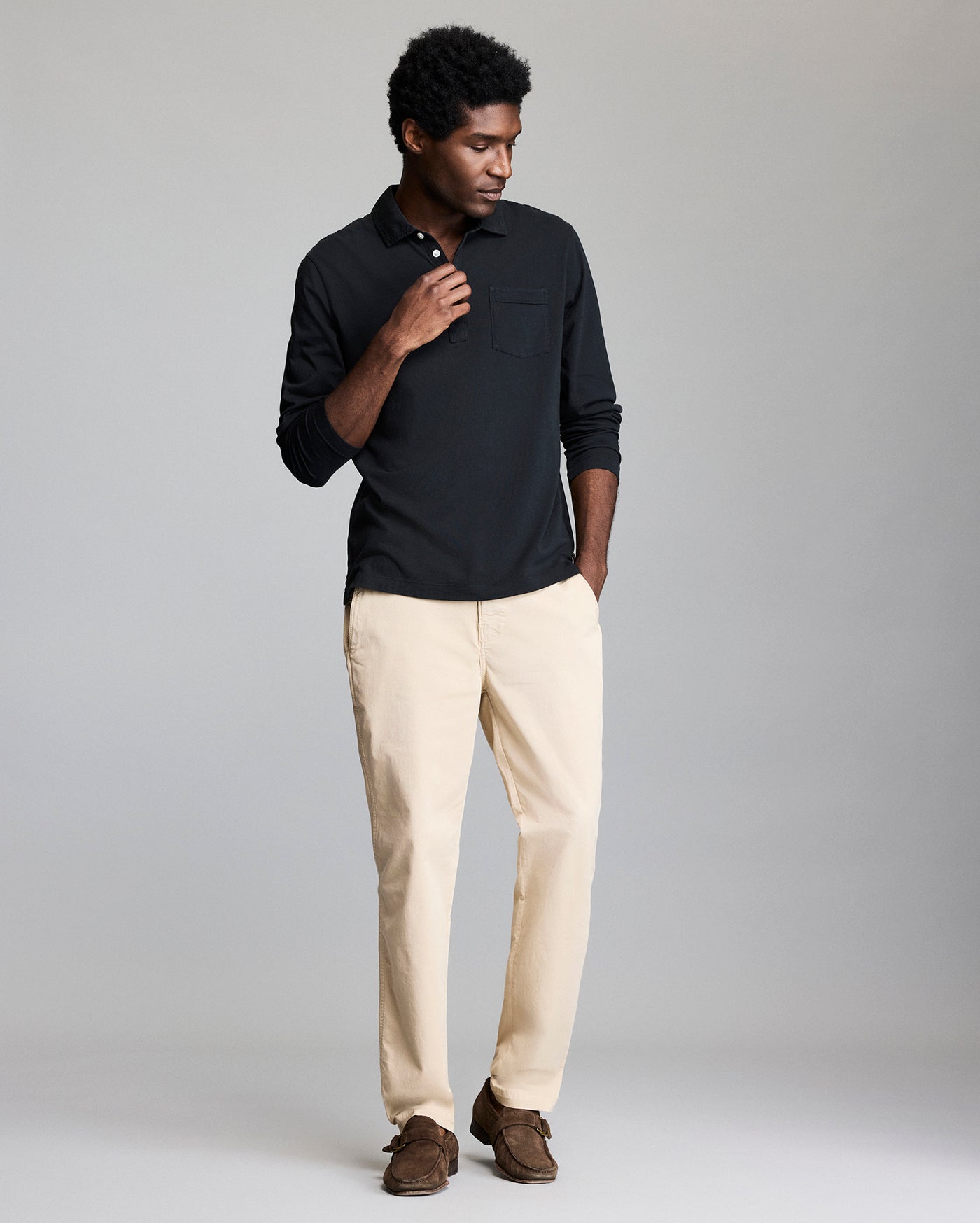 A man stands against a plain background wearing a long-sleeve black polo shirt, light beige pants, and brown shoes. He has one hand near his collar and the other in his pocket, with a relaxed, casual pose.
