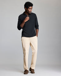 A man stands against a plain background wearing a long-sleeve black polo shirt, light beige pants, and brown shoes. He has one hand near his collar and the other in his pocket, with a relaxed, casual pose.