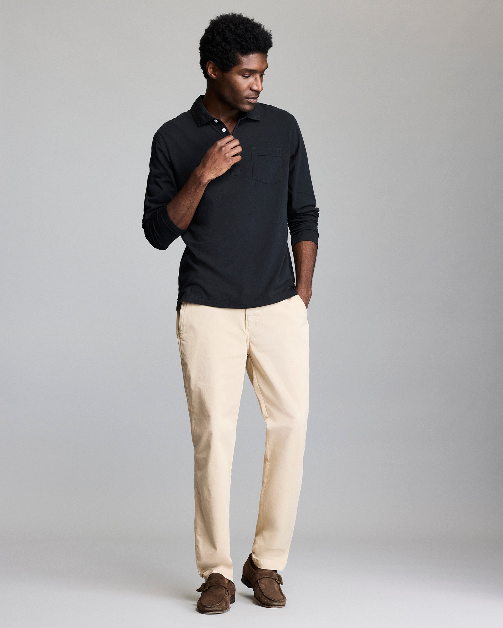 A man stands against a plain background wearing a long-sleeve black polo shirt, light beige pants, and brown shoes. He has one hand near his collar and the other in his pocket, with a relaxed, casual pose.