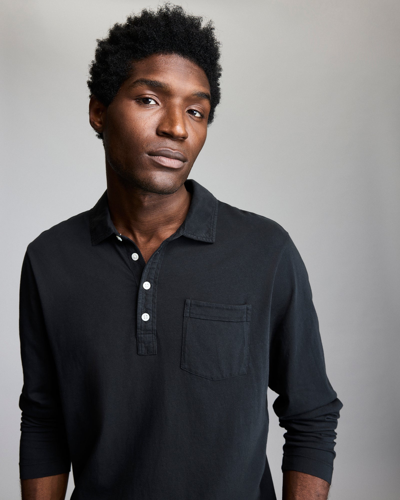 A man with short curly hair, wearing a black long-sleeve polo shirt with a chest pocket, stands against a plain light gray background, looking slightly to the side with a neutral expression.