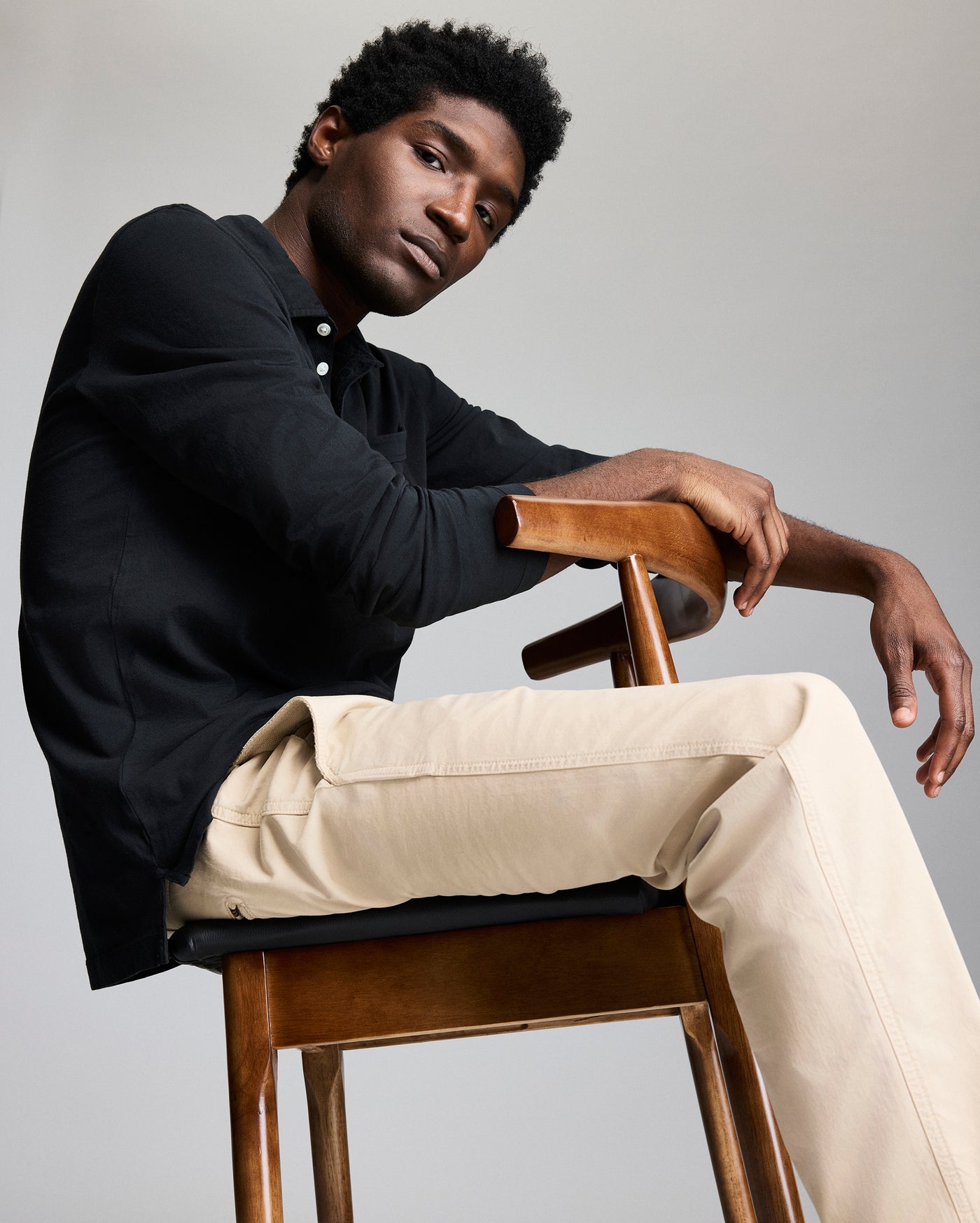 A man with short curly hair sits sideways on a wooden chair, resting his arms on the chair back. He wears a black long-sleeve shirt and beige pants, looking confidently at the camera against a plain background.
