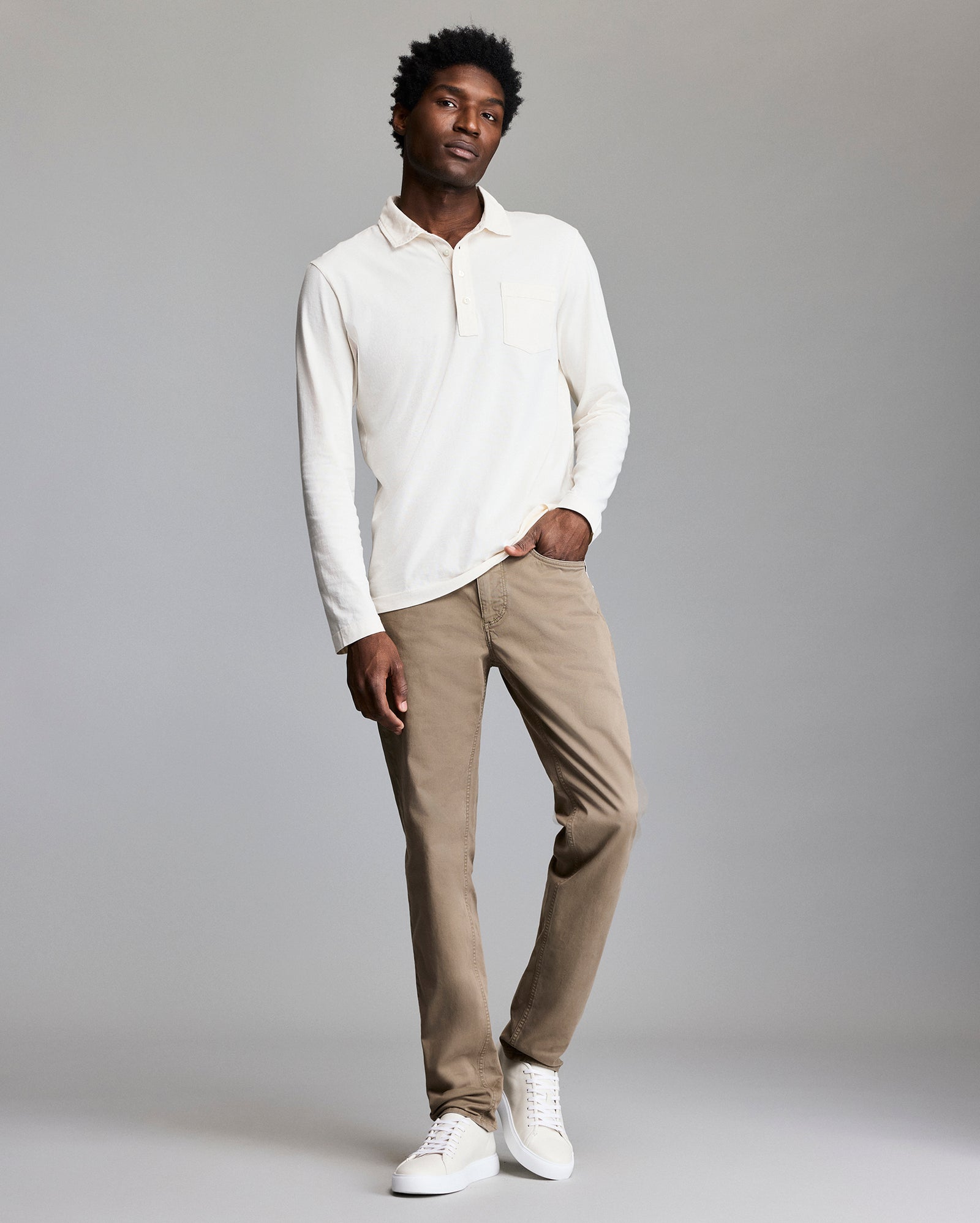 A man stands against a light gray background wearing a cream long-sleeve shirt, light brown pants, and white sneakers. He has one hand in his pocket and a relaxed expression.