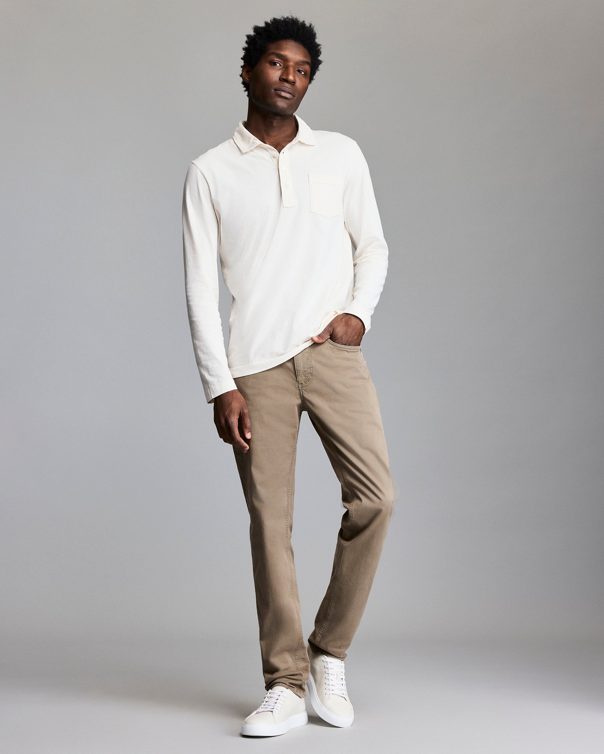 A man stands against a light gray background wearing a cream long-sleeve shirt, light brown pants, and white sneakers. He has one hand in his pocket and a relaxed expression.