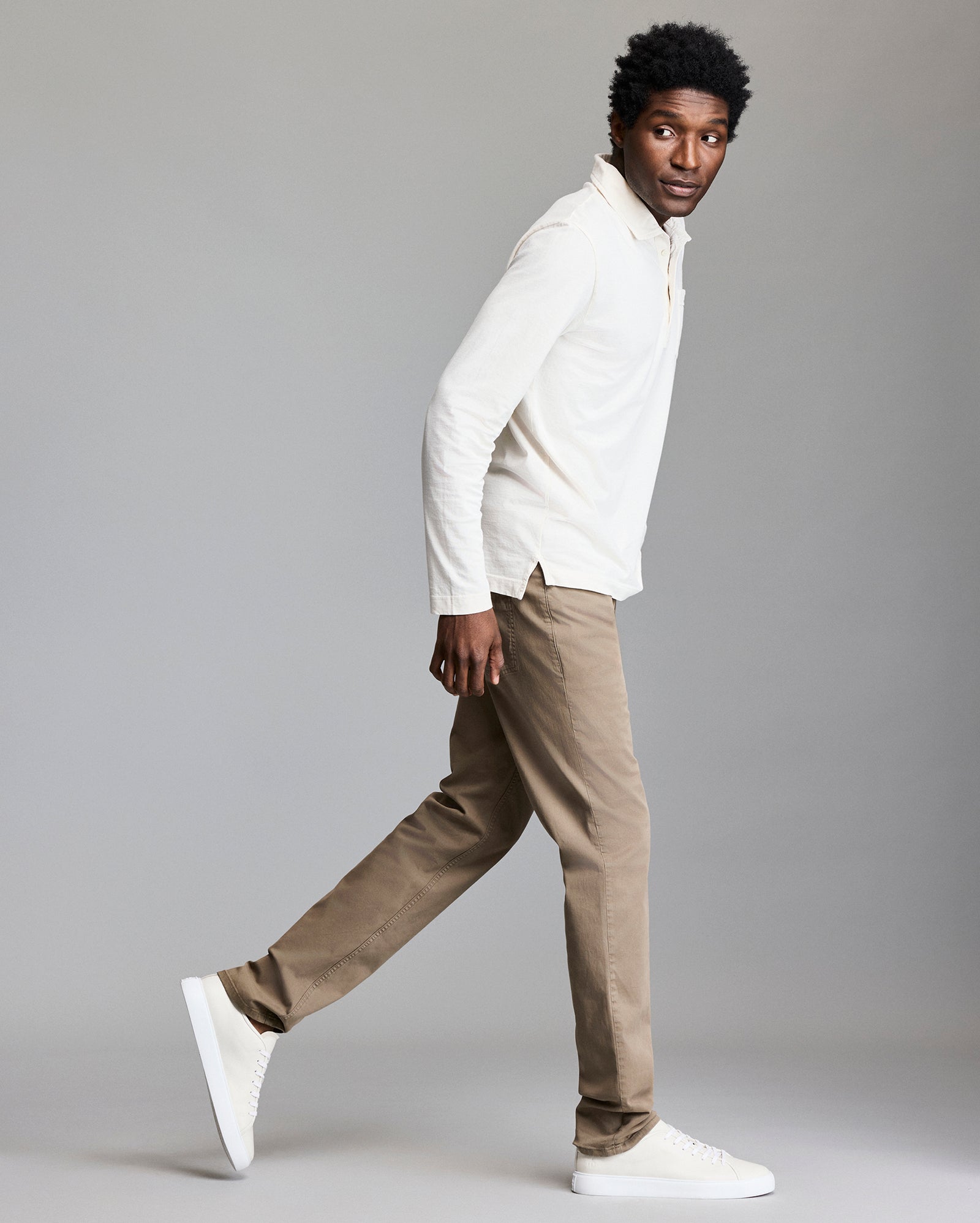 A man wearing a white long-sleeve shirt, light brown pants, and white sneakers is walking in profile against a plain gray background. He is looking slightly over his shoulder.