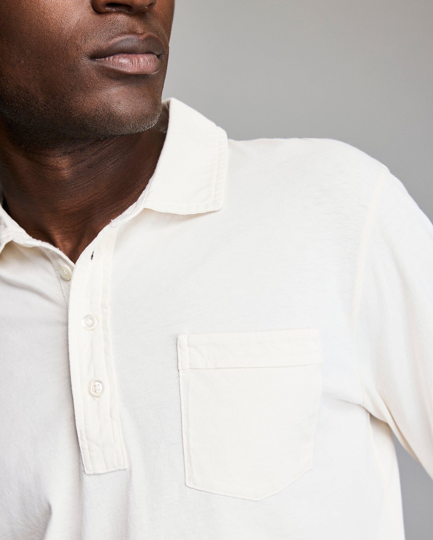 A close-up of a person wearing an off-white collared shirt with a front pocket, featuring a button placket. The lower half of their face and upper torso are visible, against a plain background.