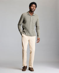 A man stands against a plain gray background wearing a sage green long-sleeve polo shirt, beige pants, and brown loafers. He looks relaxed with one hand in his pocket.