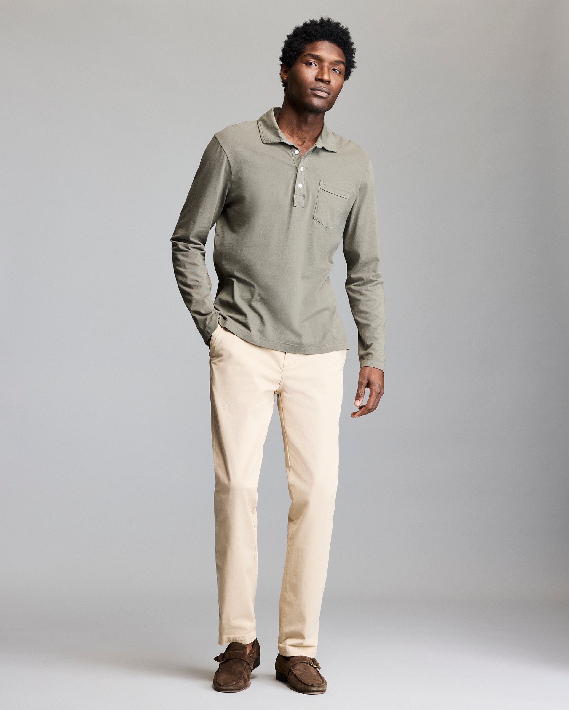 A man stands against a plain gray background wearing a sage green long-sleeve polo shirt, beige pants, and brown loafers. He looks relaxed with one hand in his pocket.