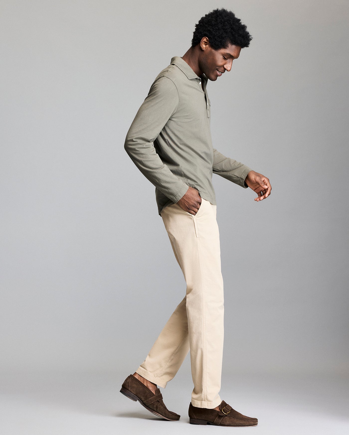 A man in a light green long-sleeve shirt, beige pants, and brown loafers walks to the right against a plain gray background, looking down with a slight smile.