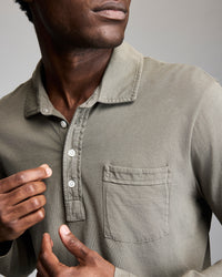 A person wearing an olive green button-up shirt with a chest pocket, partially buttoned, adjusting the collar with one hand against a plain background.