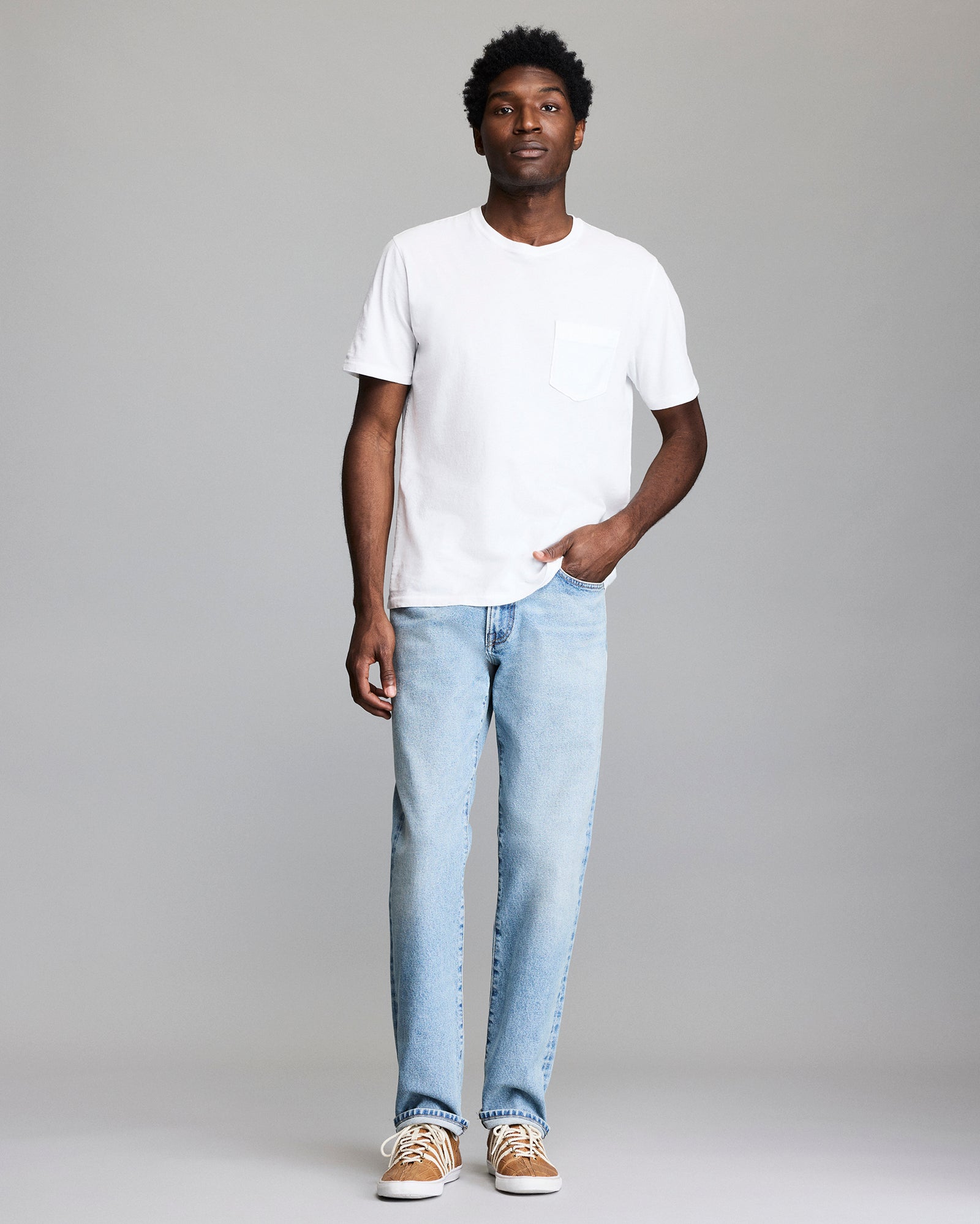 A man stands against a plain background wearing a white short-sleeve T-shirt with a chest pocket, light blue jeans, and tan sneakers. He has one hand in his pocket and a neutral expression.