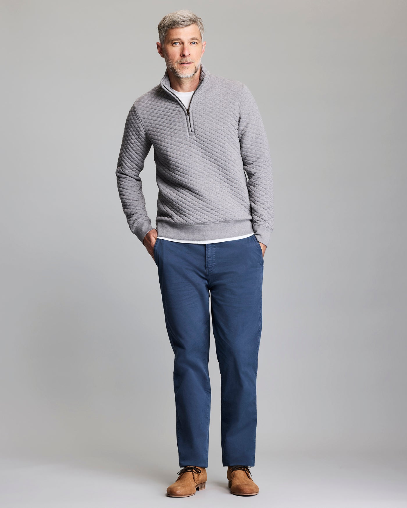 A man with gray hair, wearing a light gray quilted zip-up sweater, white shirt, blue pants, and brown shoes, stands with his hands in his pockets against a plain gray background.