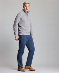 A man with short gray hair wears a light gray quilted pullover, blue pants, and brown dress shoes, standing against a plain gray background and looking slightly to the side.