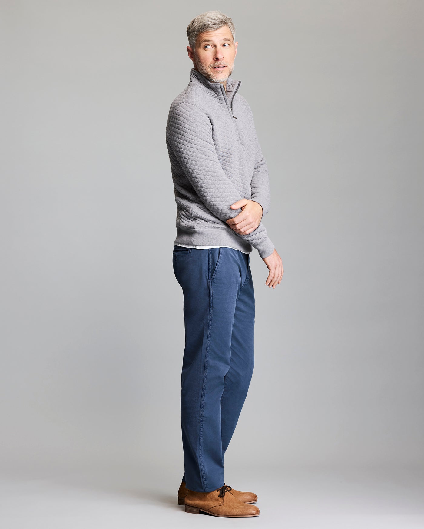 A man with short gray hair stands sideways, wearing a light gray quilted pullover, blue pants, and brown shoes. He looks slightly over his shoulder against a plain light gray background.