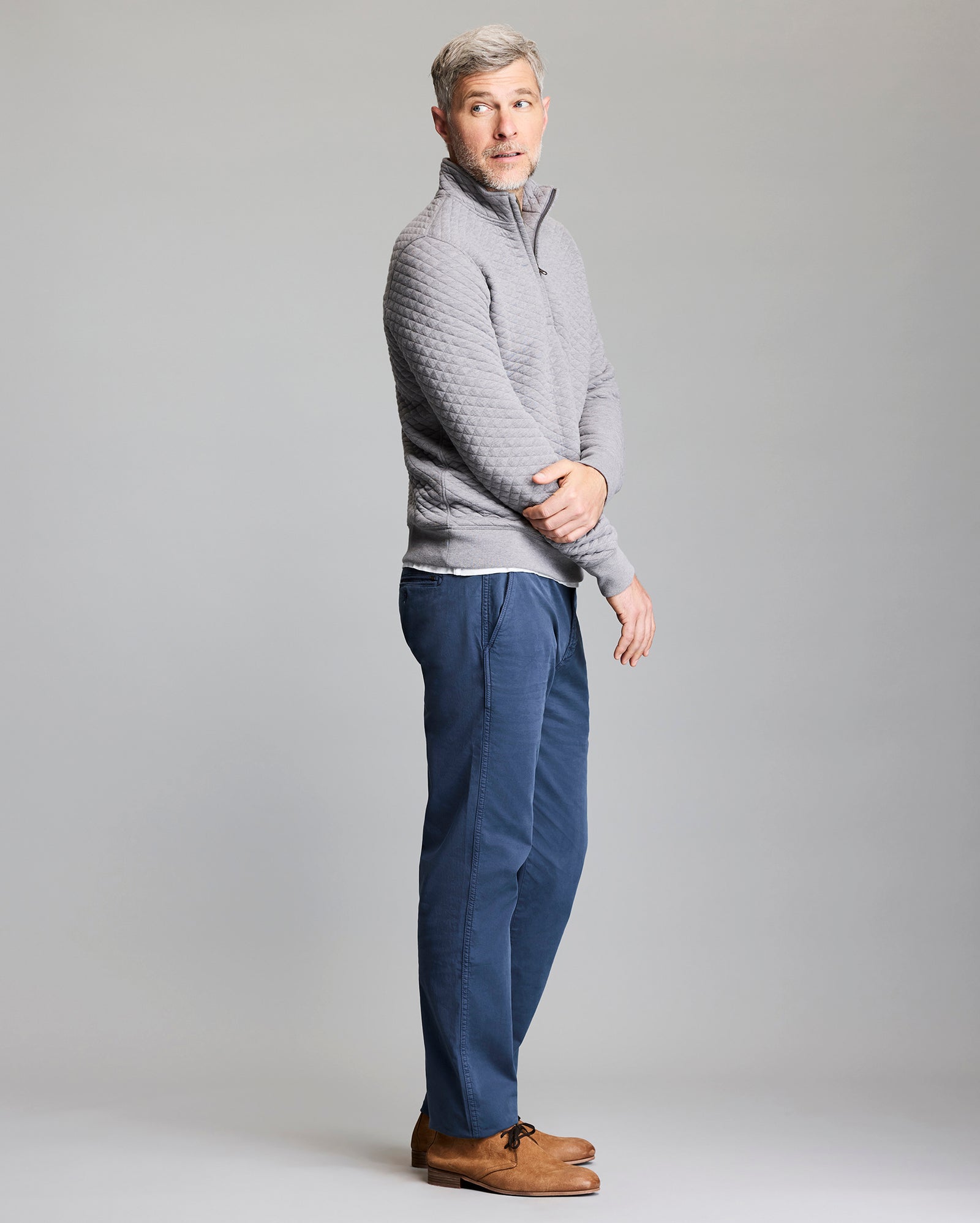 A man with short gray hair stands sideways, wearing a light gray quilted pullover, blue pants, and brown shoes. He looks slightly over his shoulder against a plain light gray background.