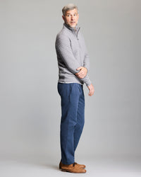 A man with short gray hair stands sideways, wearing a light gray quilted pullover, blue pants, and brown shoes. He looks slightly over his shoulder against a plain light gray background.