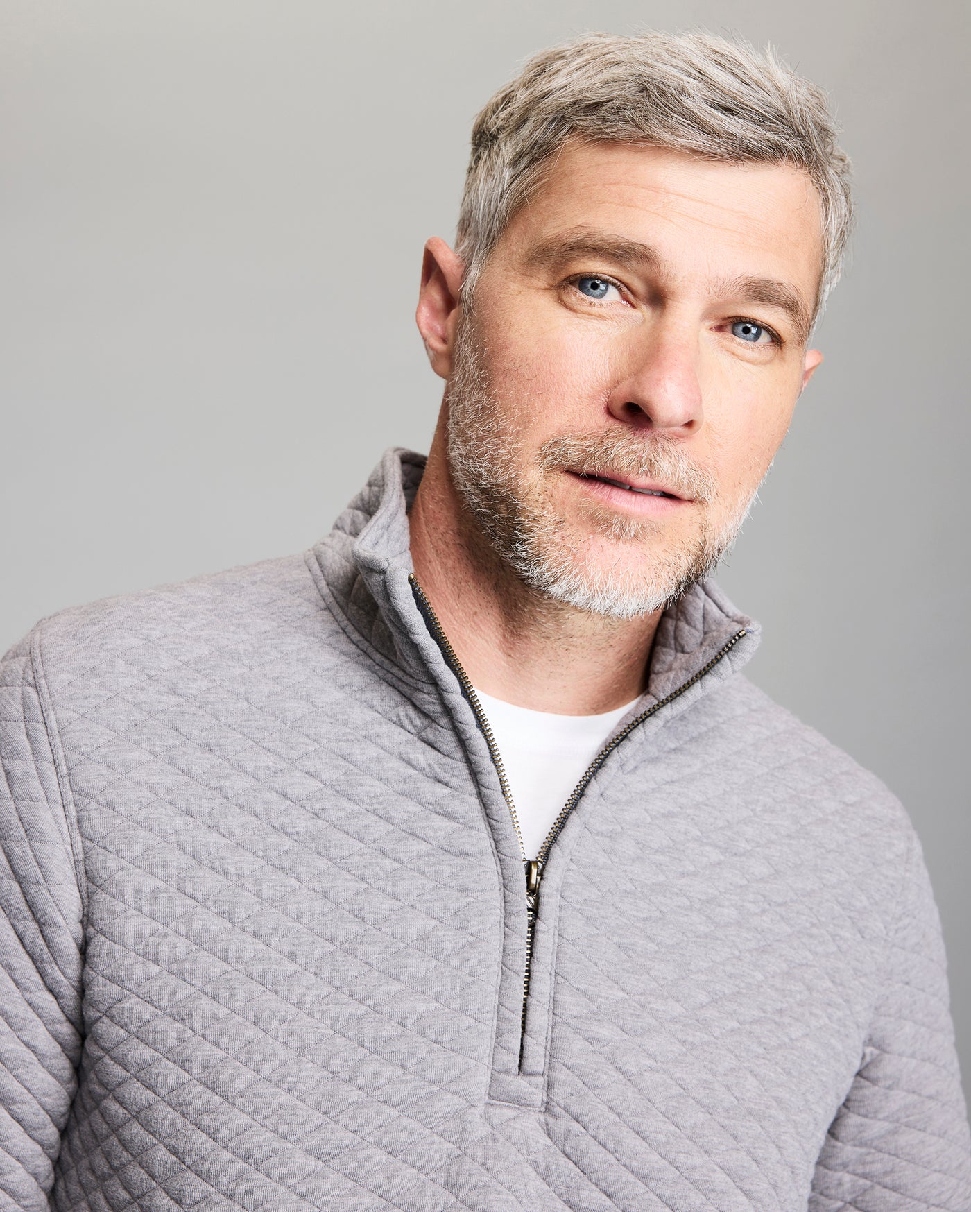 A man with short gray hair and blue eyes is wearing a light gray, quilted zip-up sweater over a white shirt. He has a short beard and is looking slightly to the right, standing in front of a plain gray background.