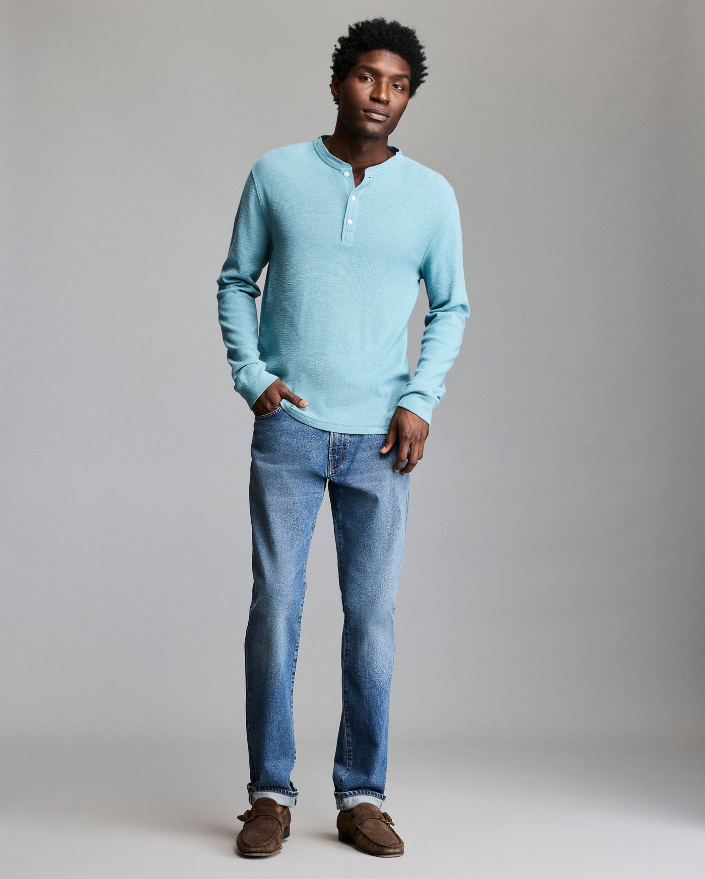 A man stands against a plain background wearing a light blue long-sleeve henley shirt, blue jeans, and brown shoes, with one hand in his pocket and a relaxed expression.