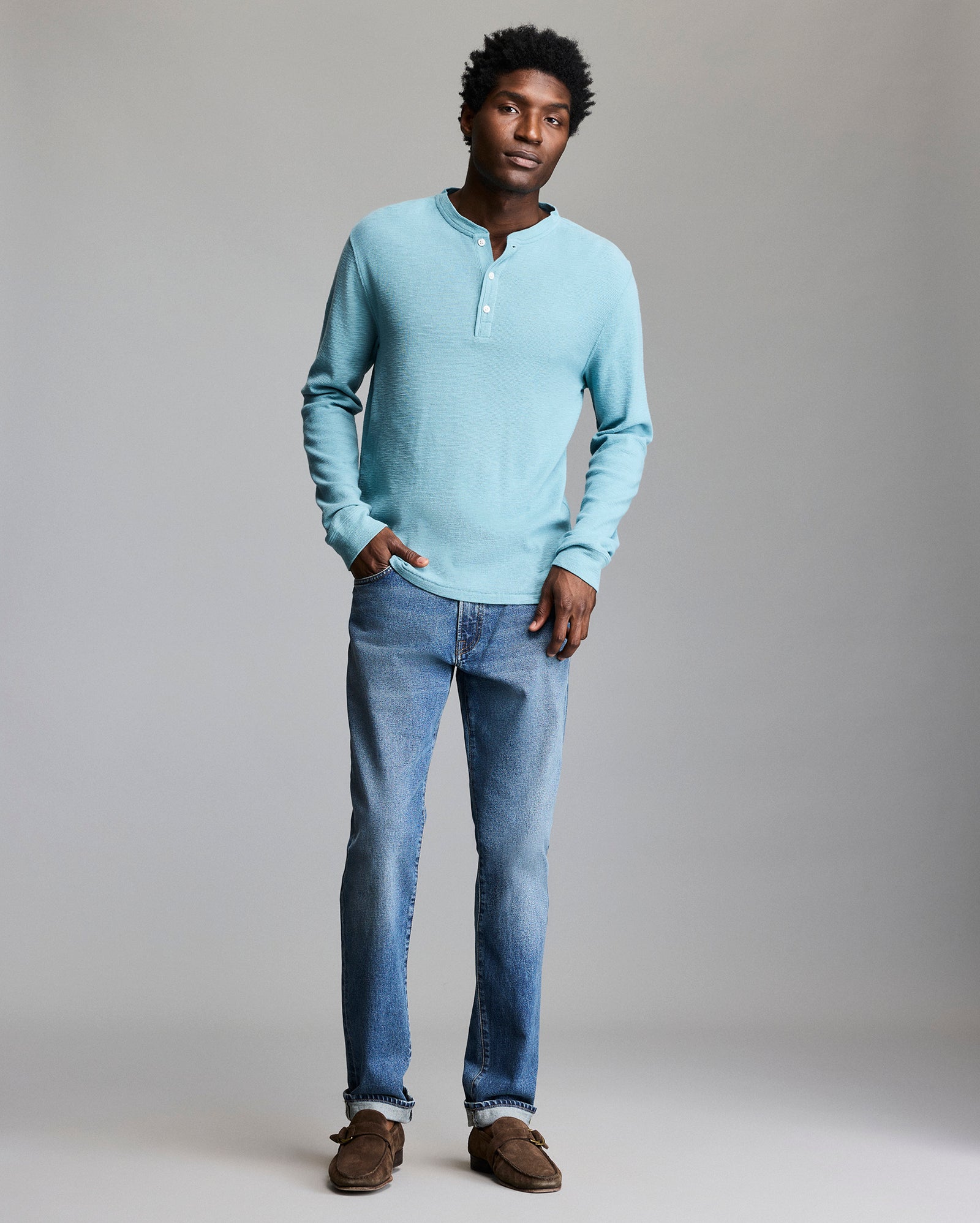 A man stands against a plain background wearing a light blue long-sleeve henley shirt, blue jeans, and brown shoes, with one hand in his pocket and a relaxed expression.