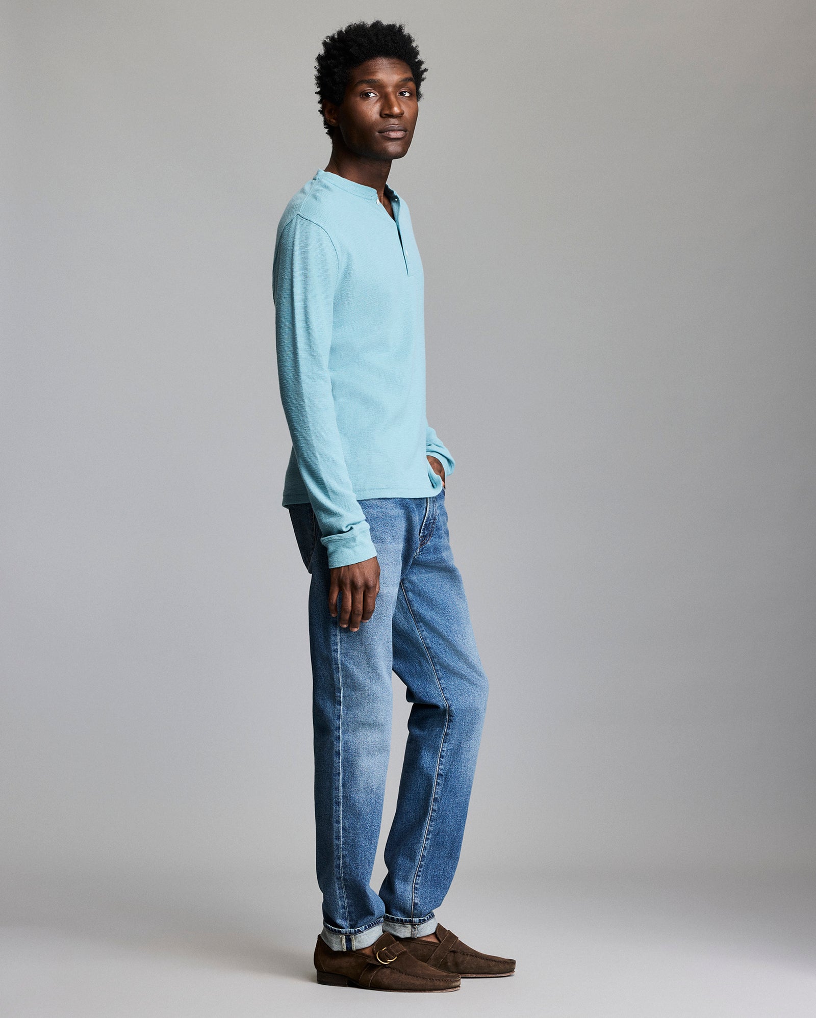A man stands in profile against a plain background, wearing a light blue Henley shirt, blue jeans, and brown suede shoes. His left hand is in his pocket and he looks calmly toward the camera.