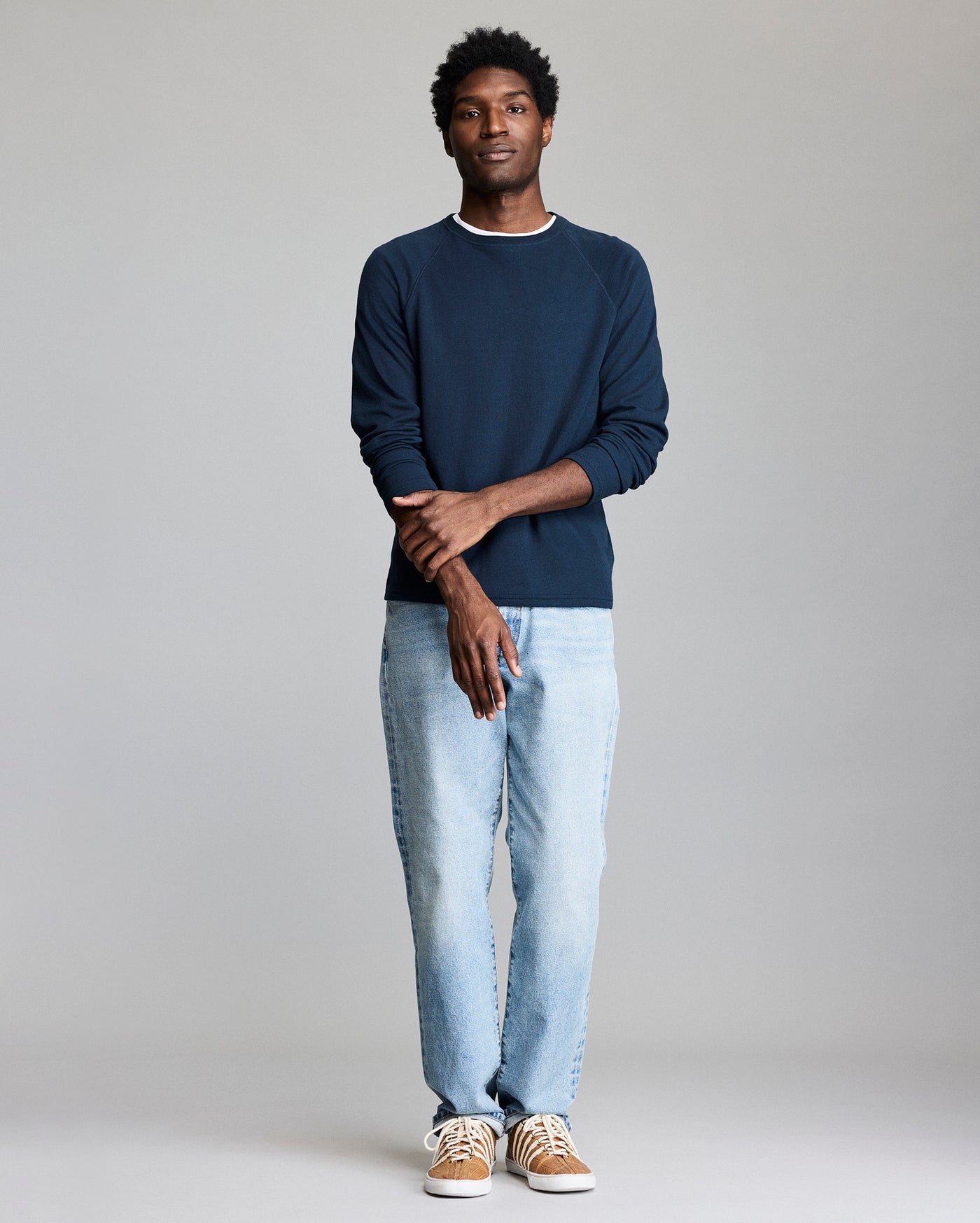 A person stands against a plain light gray background, wearing a navy blue long-sleeve shirt, light blue jeans, and tan sneakers with white laces. They stand with arms crossed and a relaxed expression.